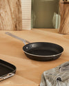 Black non-stick frying pan by Alessi with a silver handle, resting on a wooden counter beside a striped towel.