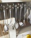 Polished stainless steel kitchen utensil set by Alessi, hanging on a metal rail above marble boards and a lemon.