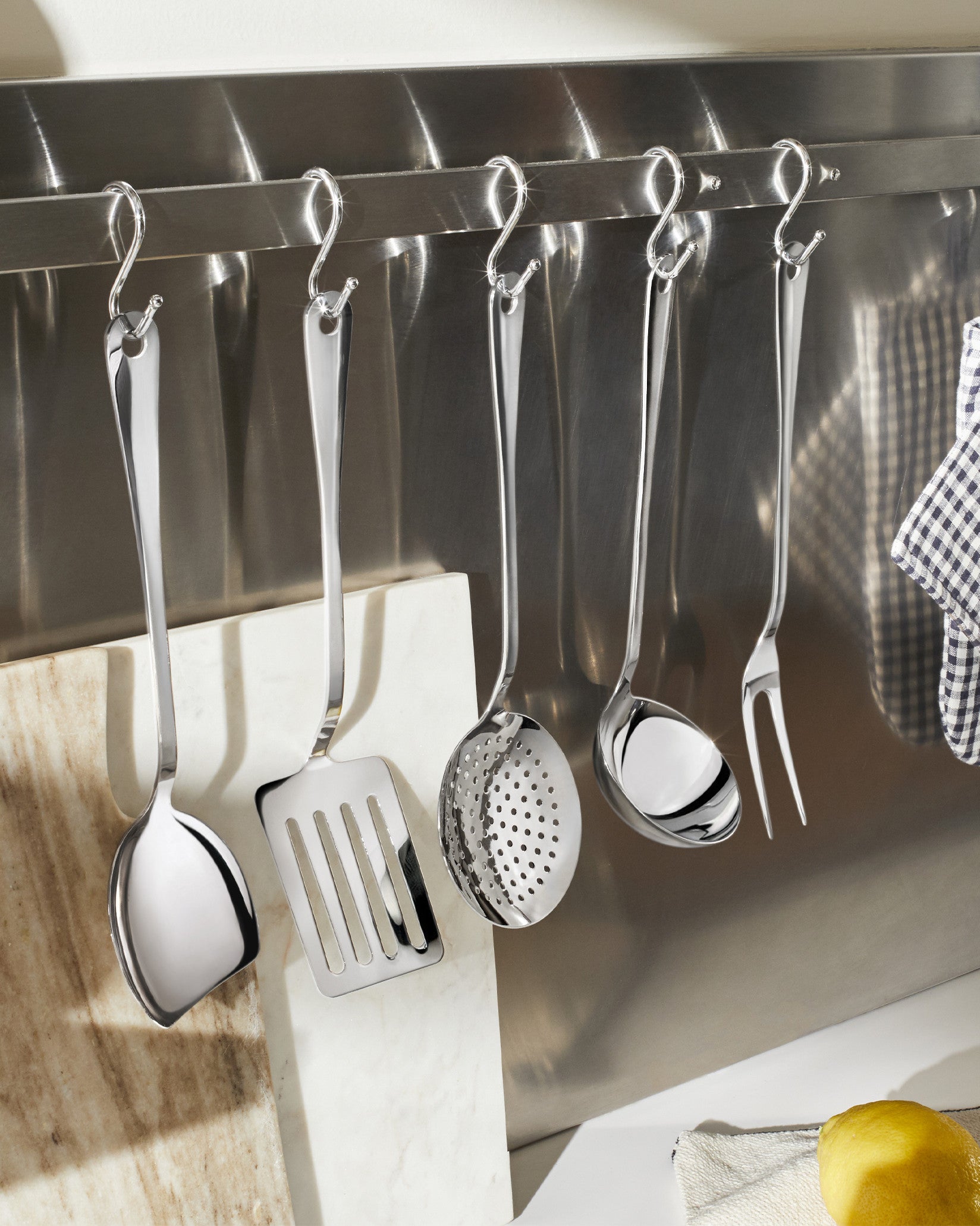Polished stainless steel kitchen utensil set by Alessi, hanging on a metal rail above marble boards and a lemon.