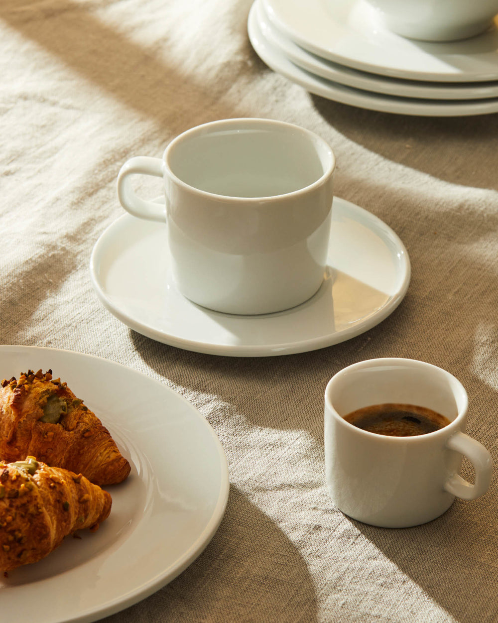 White ceramic cups and plates by Alessi, arranged with croissants and espresso on a sunlit linen tablecloth.