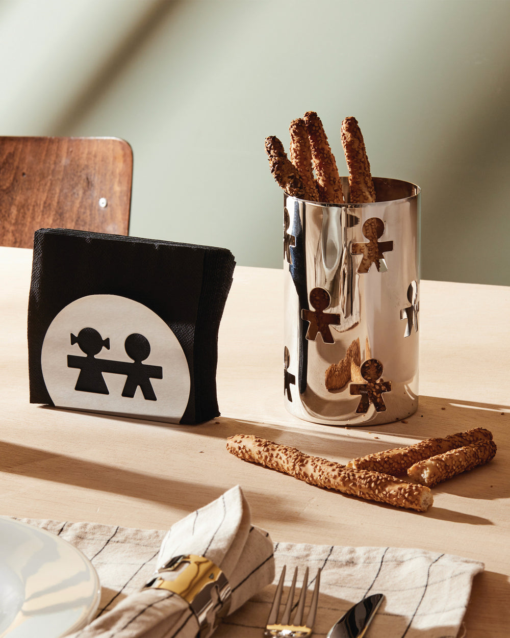 Silver metal Girotondo container by Alessi, featuring cutout figures and filled with breadsticks.