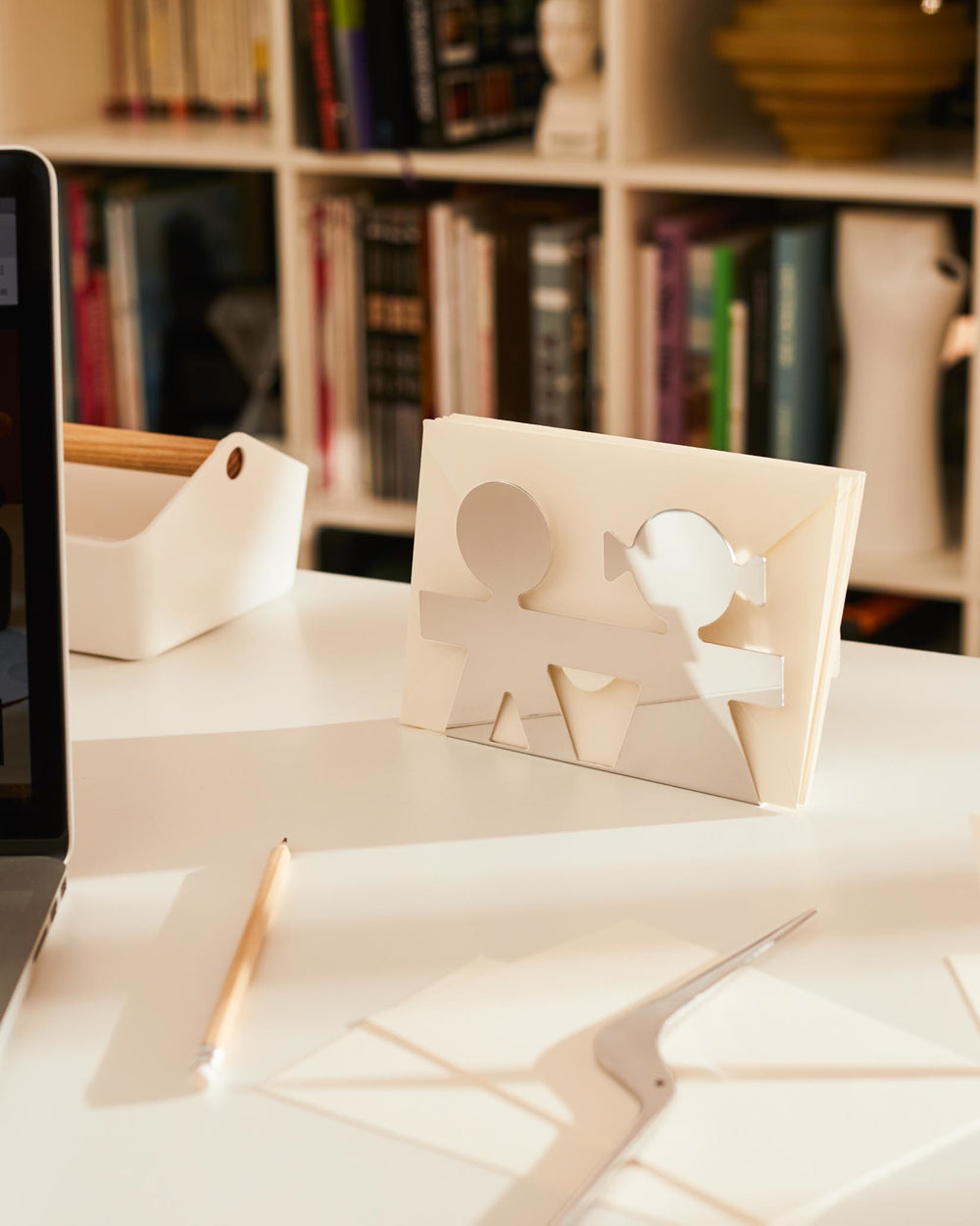 Silver-toned metal letter holder by Alessi, featuring boy and girl cut-outs, holding cream envelopes on a white desk.