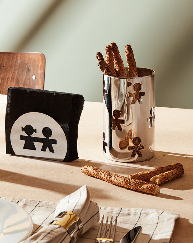 Polished silver Girotondo utensil holder by Alessi, with playful cut-out figures, holding breadsticks on a wooden table.