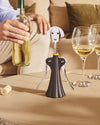 Figurative dark gray and silver Anna G. corkscrew by Alessi, with a smiling face, on a table with wine glasses.