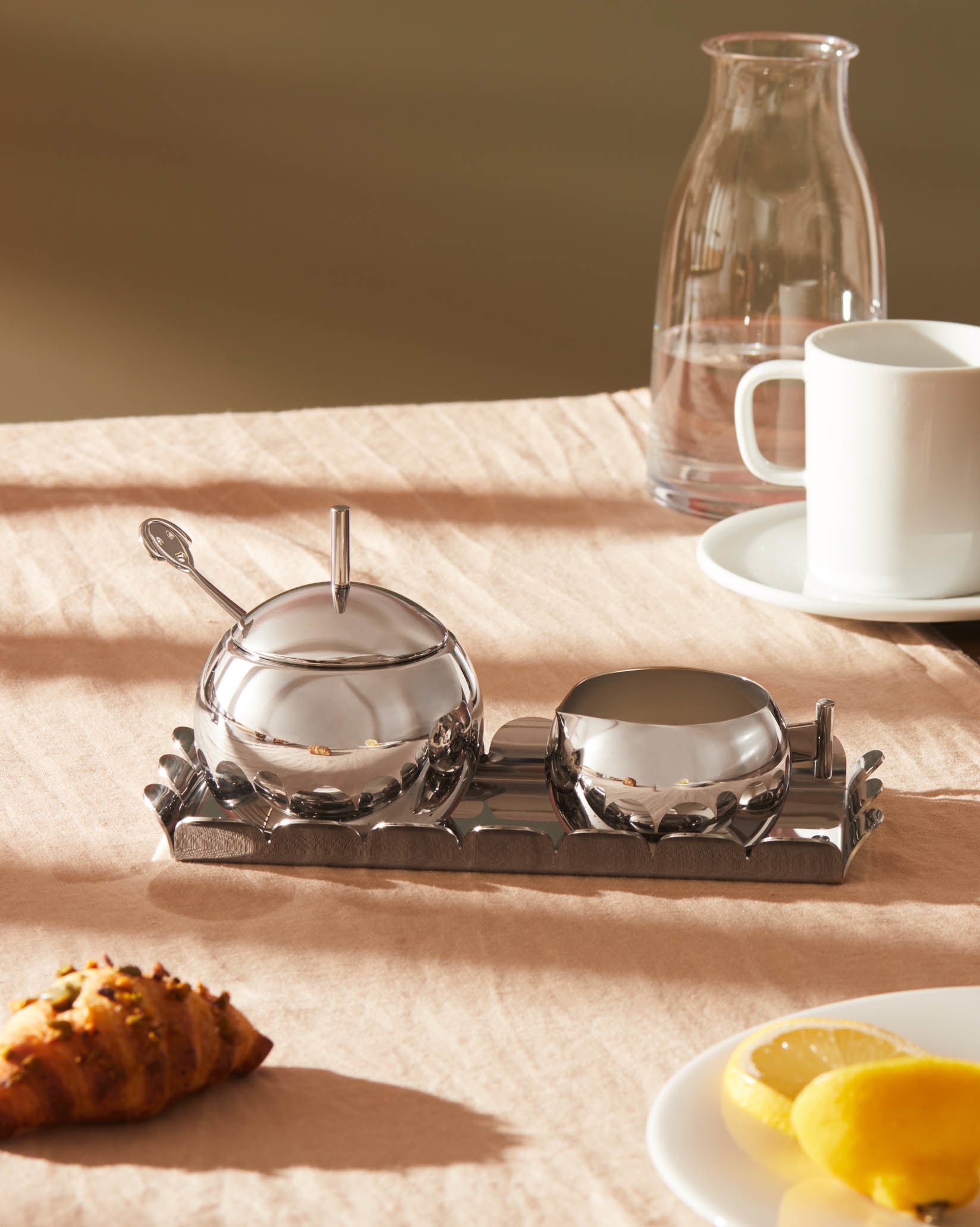 Shiny silver creamer by Alessi, a round bowl with a pouring spout, on a decorative metal tray.