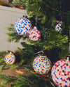 Multi-colored confetti-patterned Christmas ornament by Alessi, hanging on a green Christmas tree.