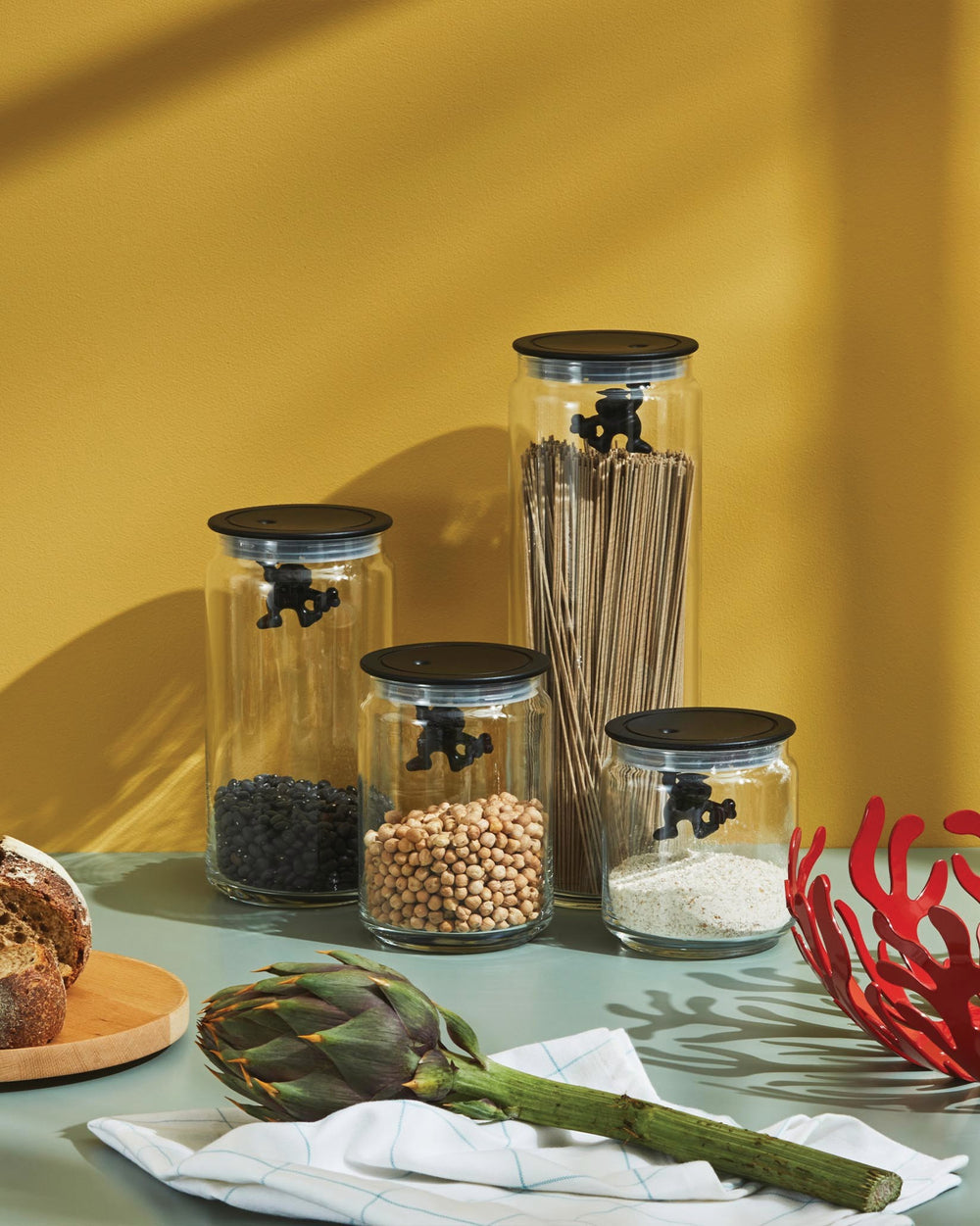 Clear glass storage jars with black lids featuring 'Gianni' a little man holding on tight by Alessi, filled with dry goods.