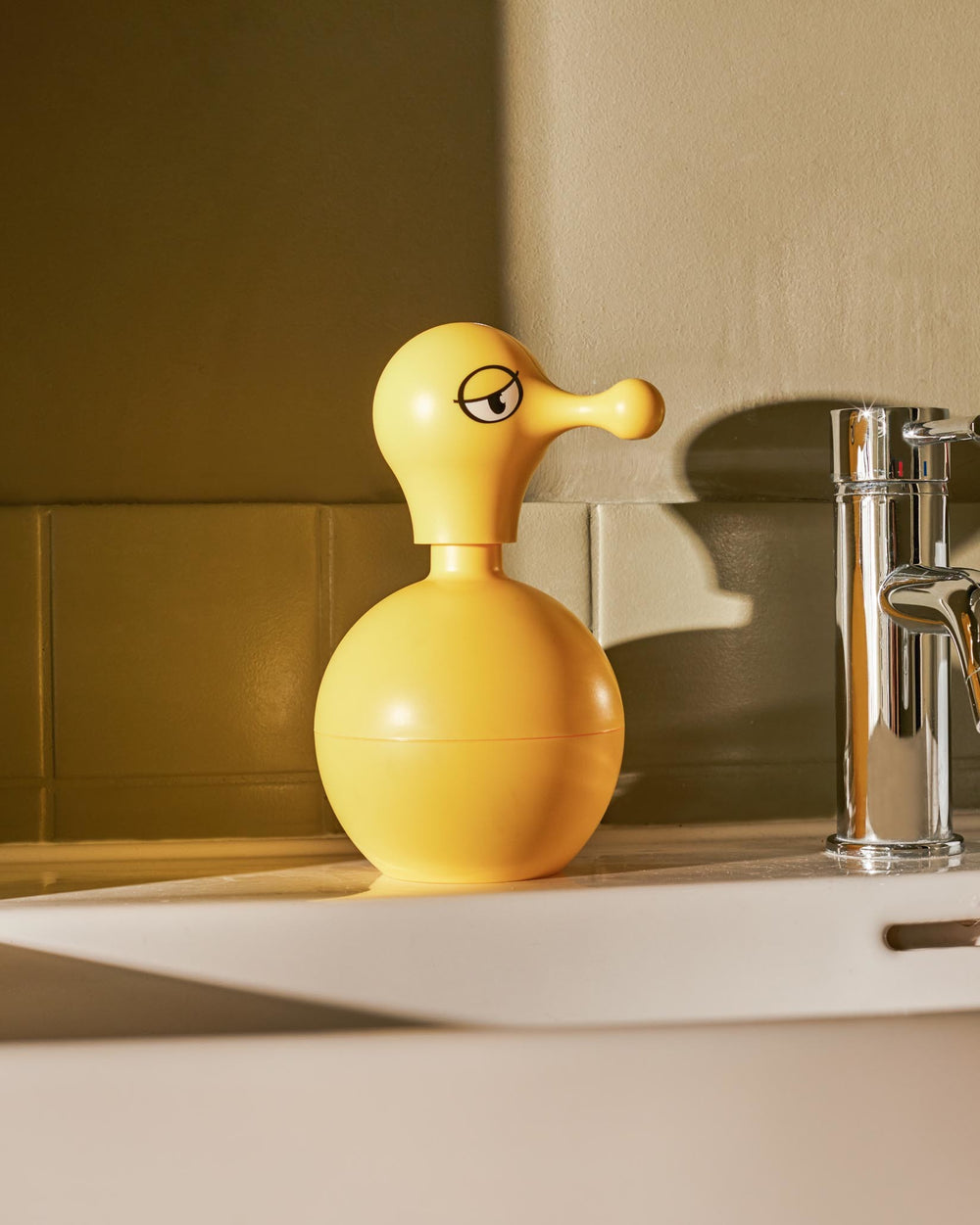 Yellow duck-shaped soap dispenser by Alessi with a whimsical cartoon eye and long beak-like pump on a bathroom sink.