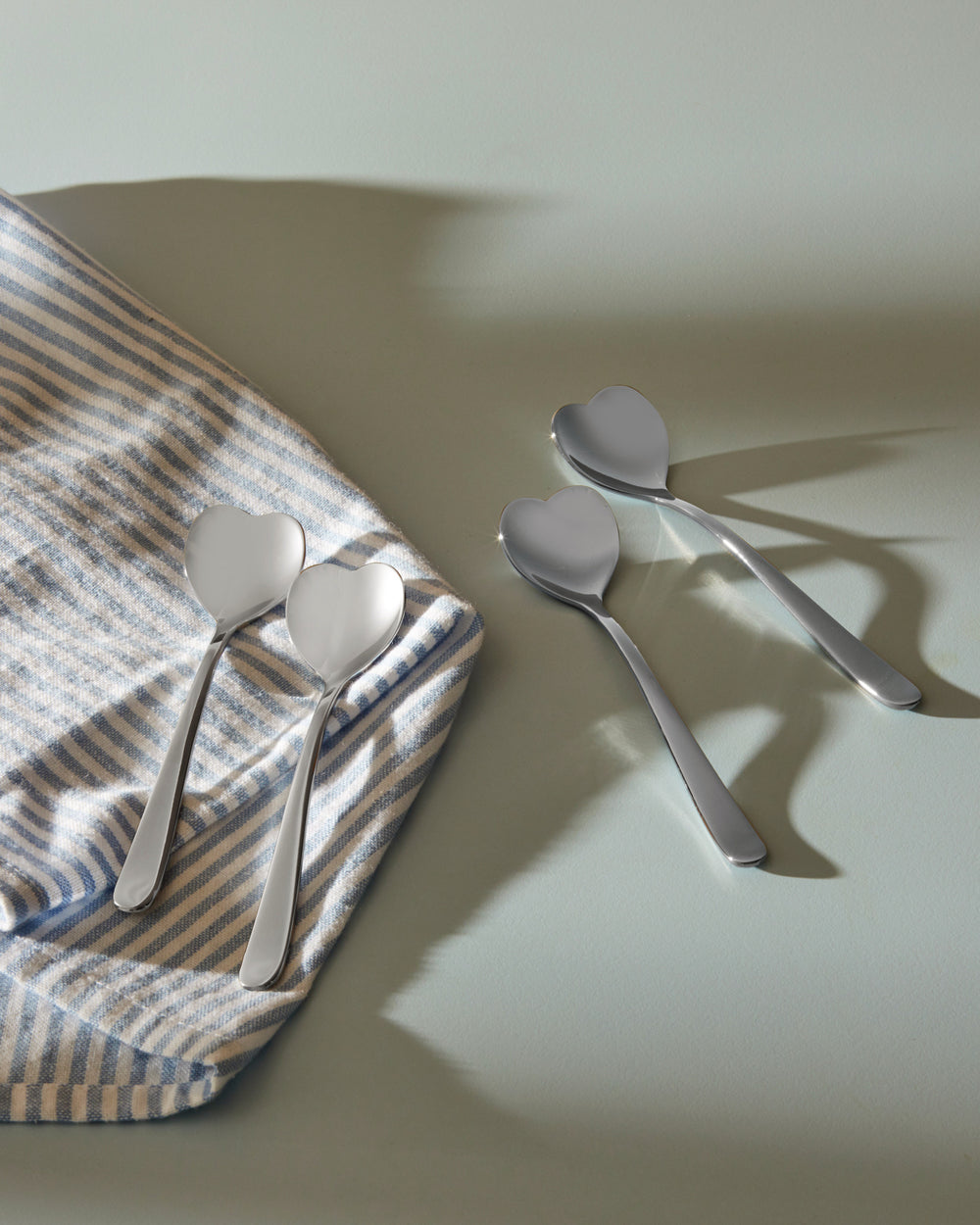 Four heart-shaped stainless steel spoons by Alessi, two on a striped blue towel and two on a light green surface.