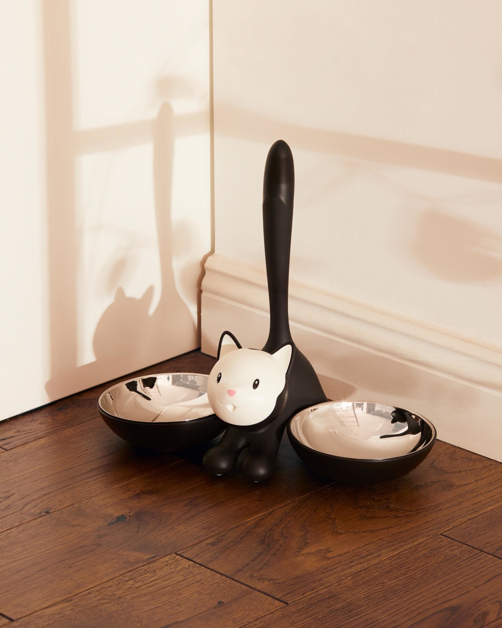 Black and white cat-shaped pet feeder with two silver bowls by Alessi, on a wooden floor.