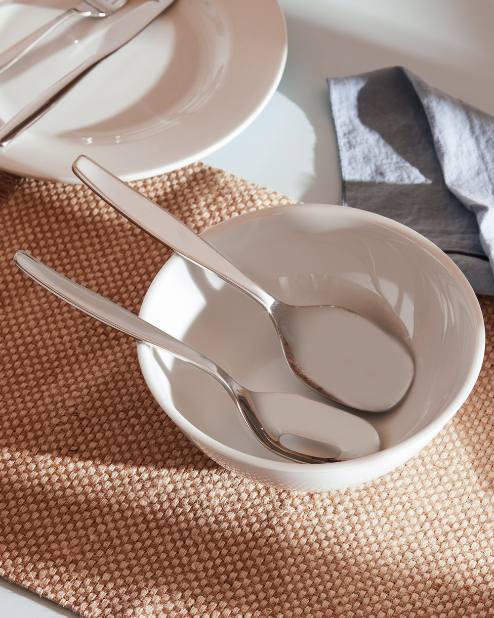 Polished silver flatware by Alessi on a woven placemat, with two spoons in a white ceramic bowl and a blue napkin.