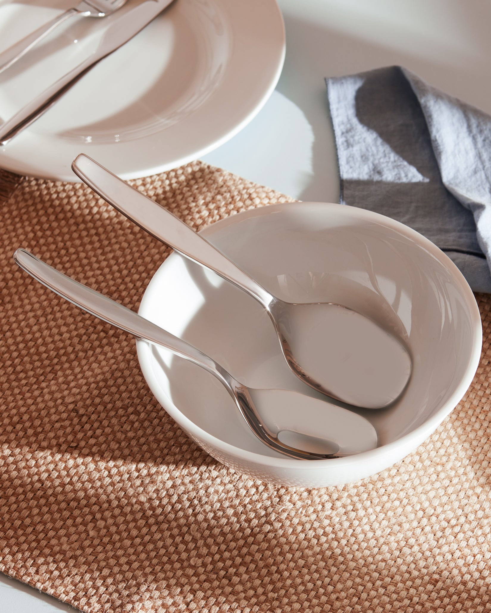 Polished silver flatware by Alessi on a woven placemat, with two spoons in a white ceramic bowl and a blue napkin.