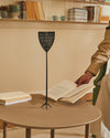 Black perforated fly swatter by Alessi with a tall handle and tripod base, standing on a light brown table.