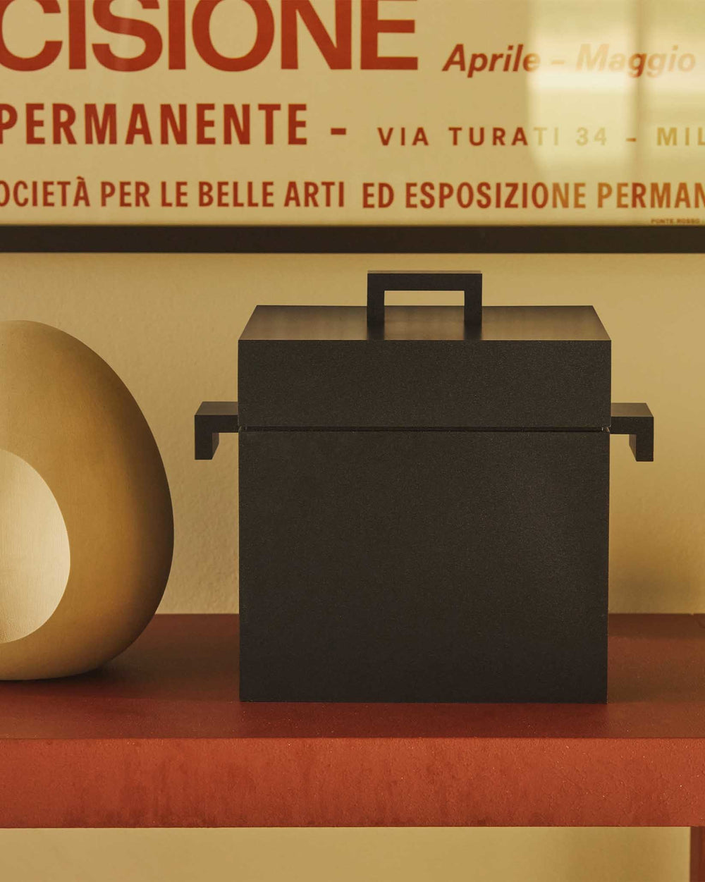 Dark cubic storage box with lid and side handles by Alessi, resting on a reddish-brown shelf.