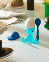 Blue translucent character egg cup by Alessi holding a dark blue spoon, with a white egg and a matching blue cap.