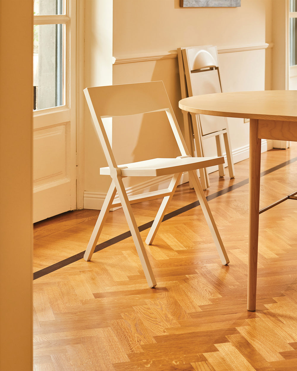 White Piana folding chair by Alessi, standing on a herringbone wood floor in a sunlit room.