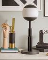 Dark bronze and white spherical table lamp by Alessi, with a fluted architectural column base, on a desk.