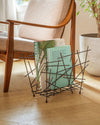 Dark metal magazine rack by Alessi with abstract intersecting rods, holding books on a wooden floor.