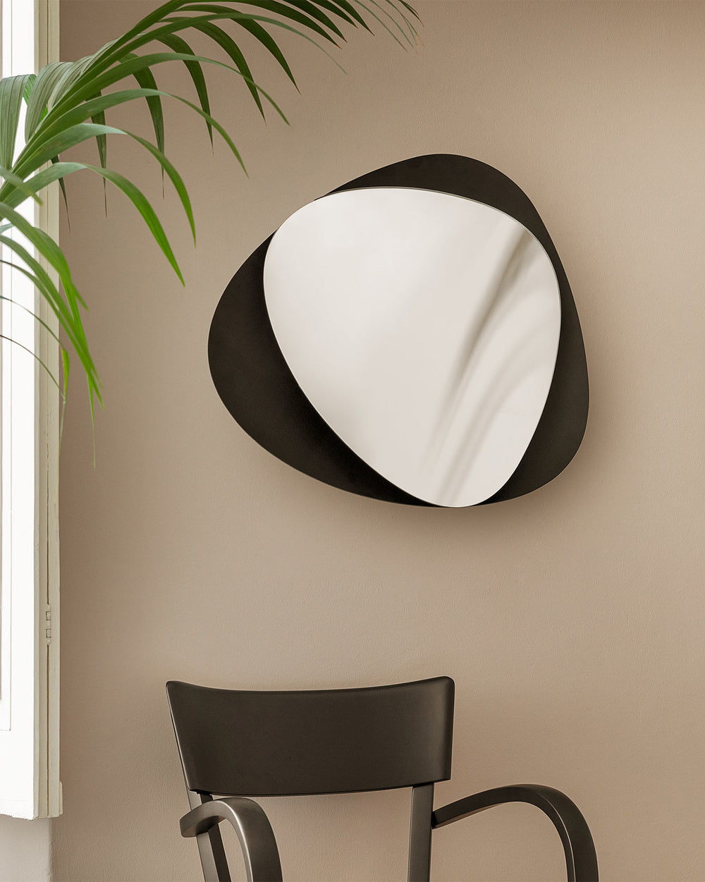 Black abstract-shaped mirror by Alessi on a beige wall, reflecting a green plant.