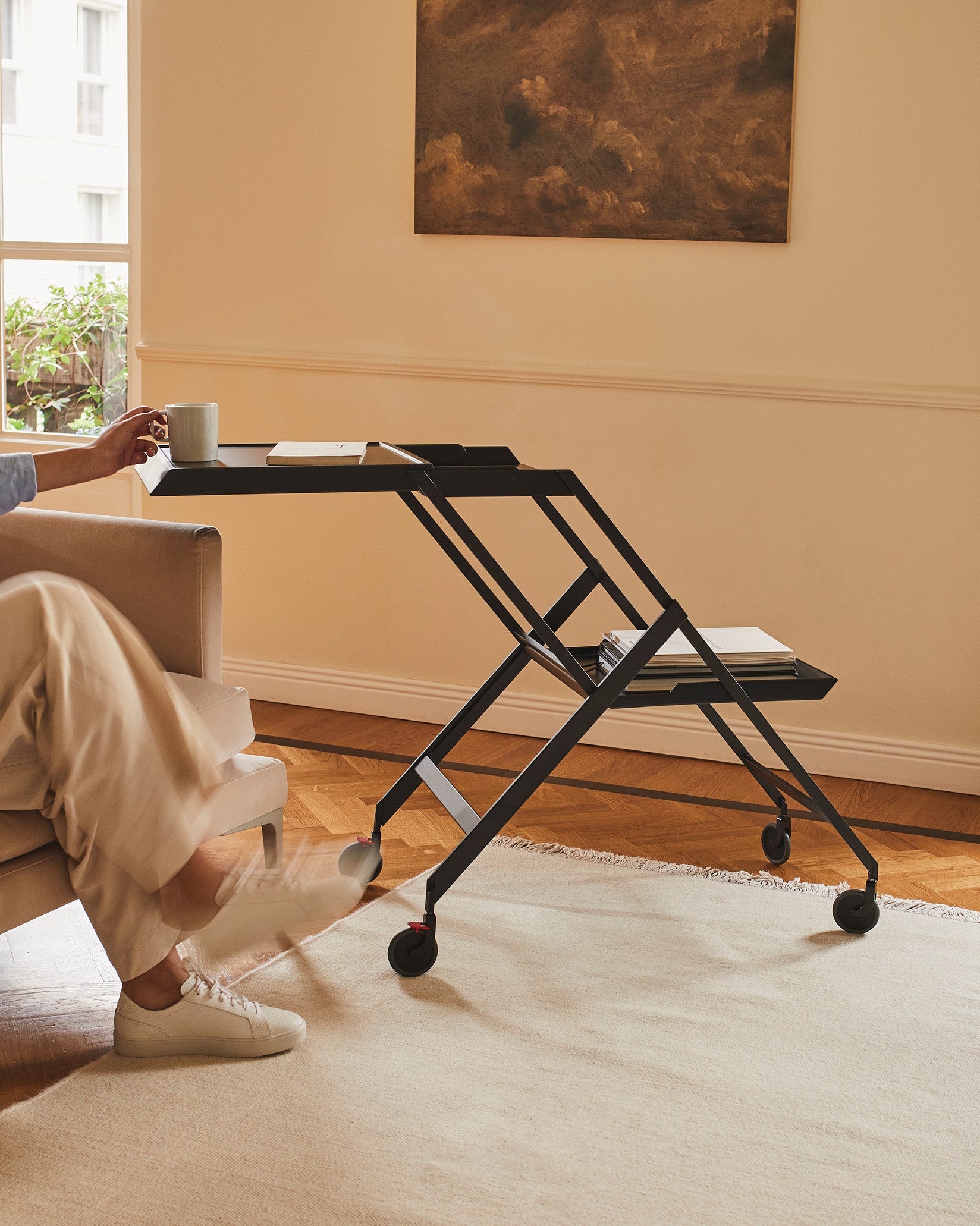 Black metal folding trolley table by Alessi, with two tiers and wheels, holding a mug and books.