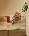 Wooden toothpick dispenser by Alessi with a silver figurine, being set by a hand on a marble countertop.