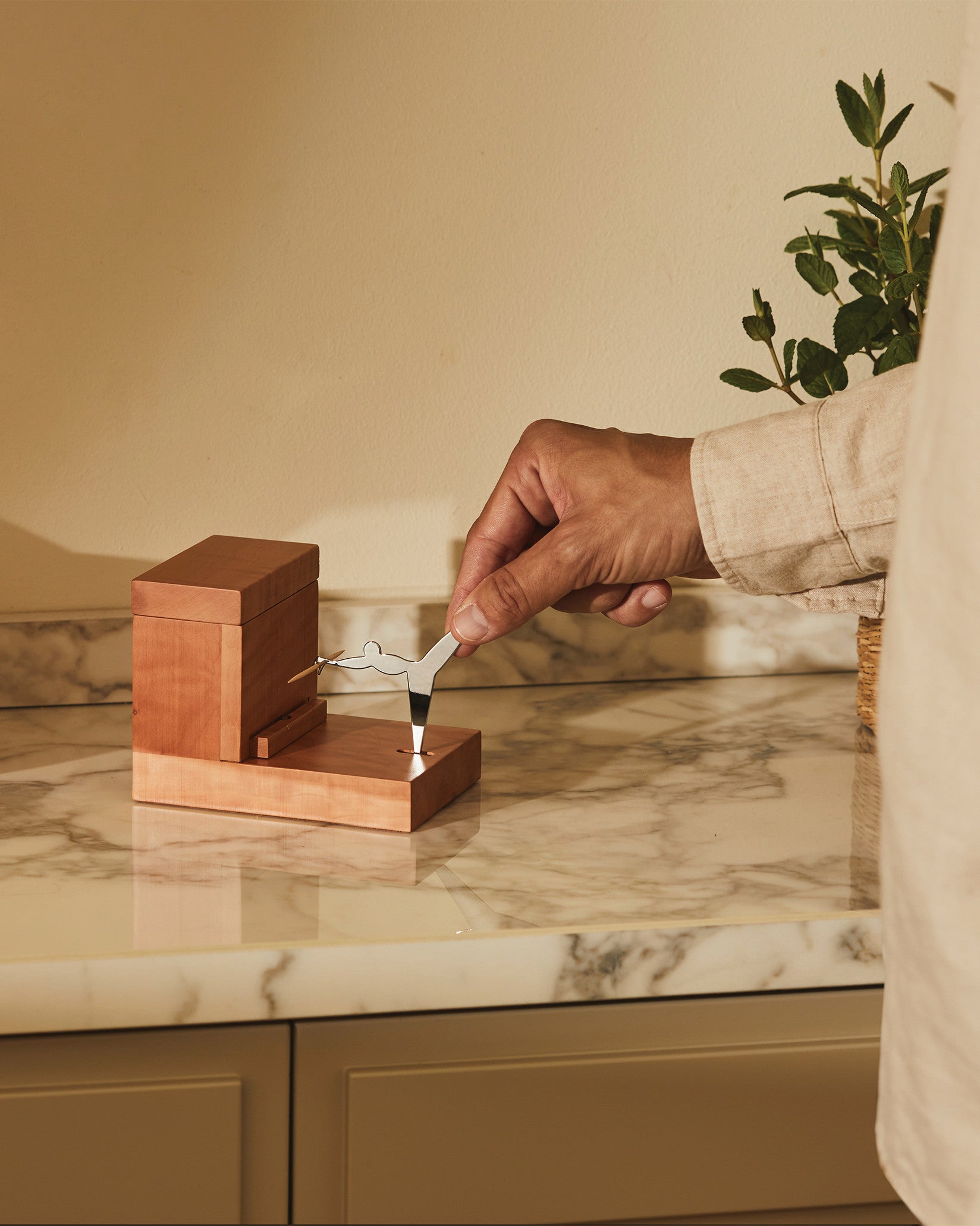 Wooden toothpick dispenser by Alessi with a silver figurine, being set by a hand on a marble countertop.