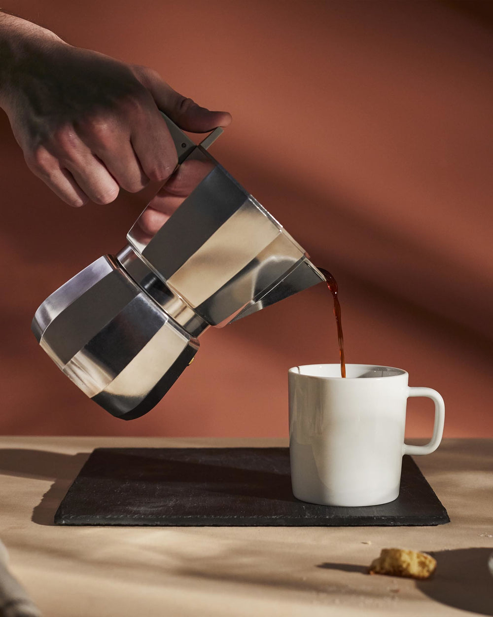 Silver metallic Moka pot by Alessi held by a hand pouring dark coffee into a white mug on a black coaster.