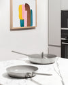 Two brushed stainless steel pans by Alessi, one with a lid, on a white marble kitchen counter.
