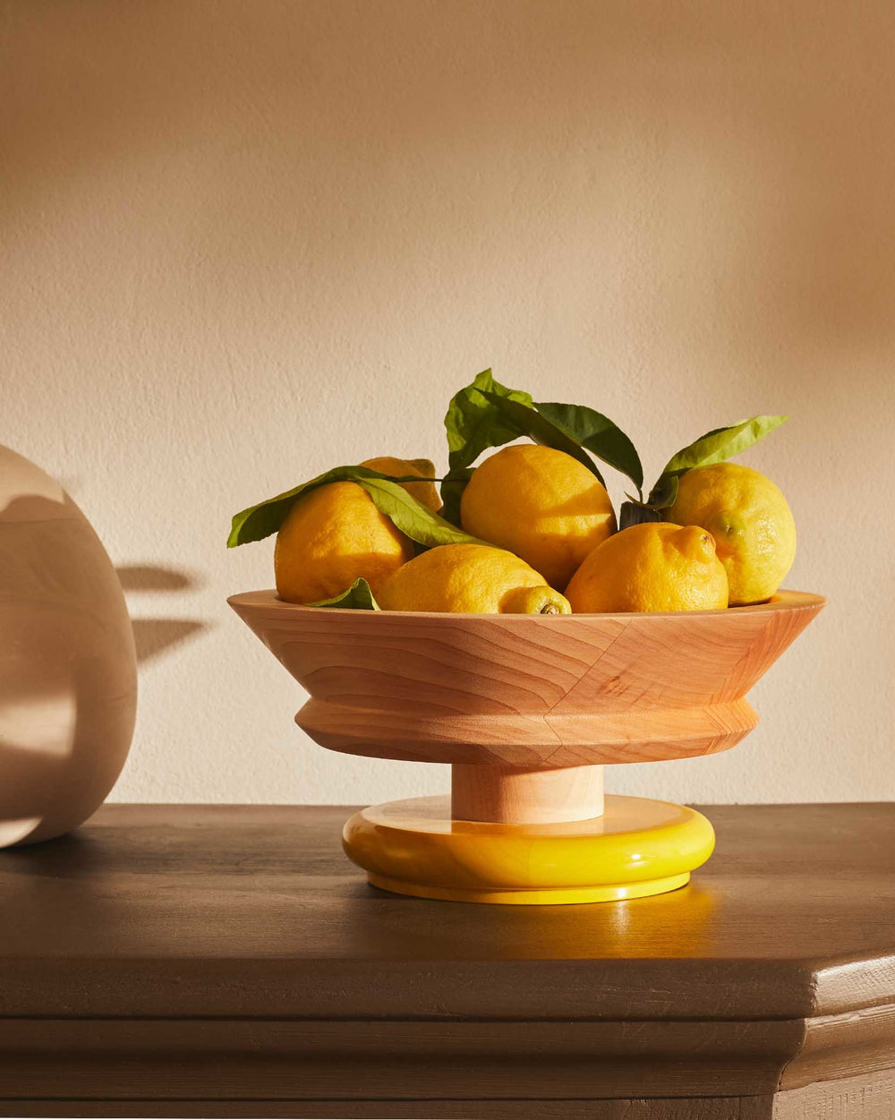 Wooden fruit bowl with a yellow base by Alessi, filled with lemons on a dark wooden surface.