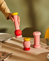Sculptural pink, red, and yellow mills by Alessi, with one held and two standing on a dining table.