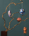 Four abstract Christmas tree ornaments by Alessi in purple, blue, red, and copper, with pixelated geometric patterns, hanging on bare branches.