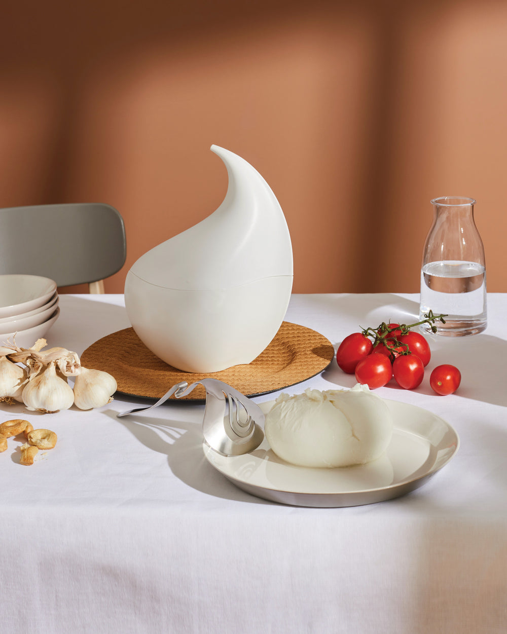 White sculptural container by Alessi, with a smooth, organic shape, styled on a table with garlic and tomatoes.