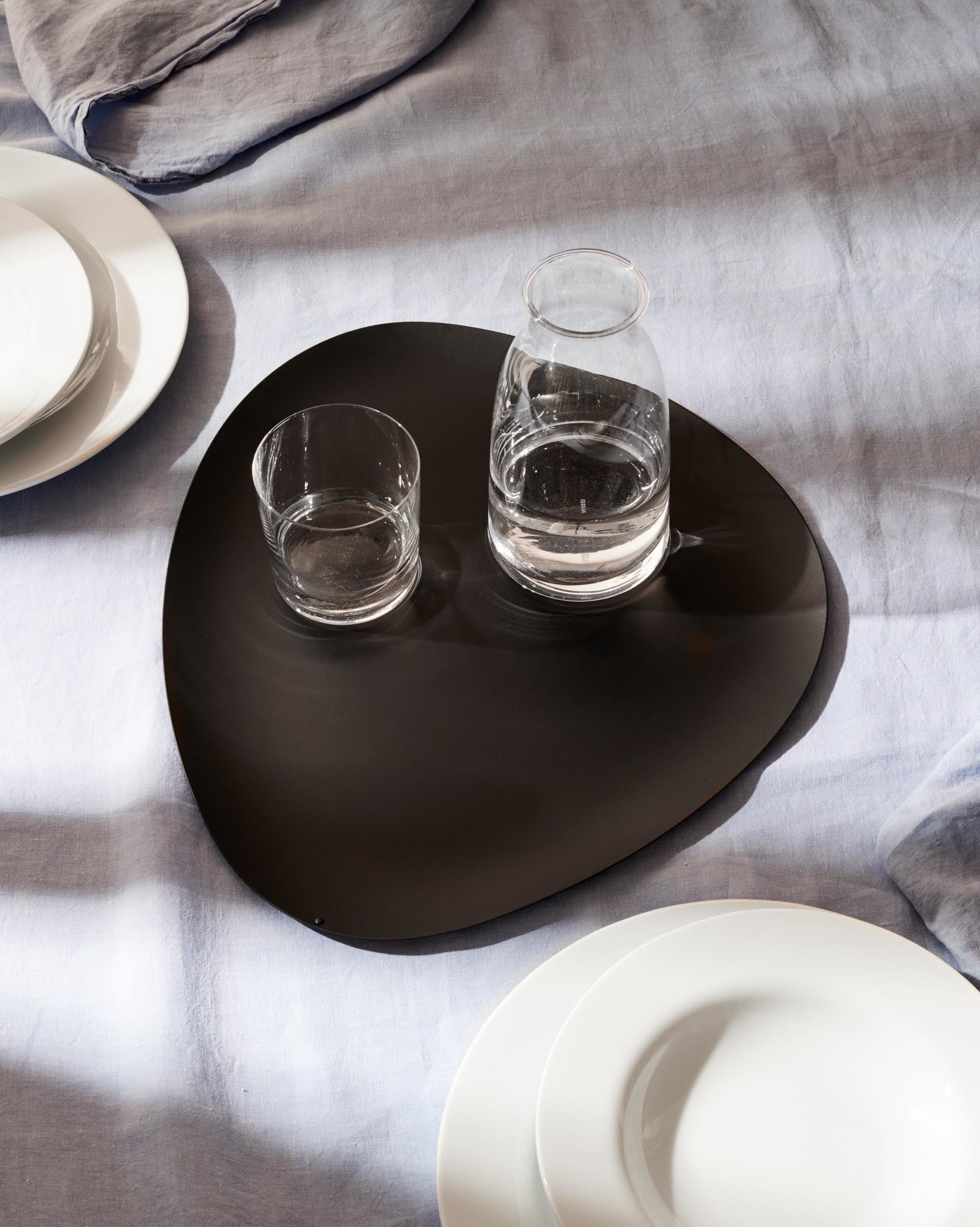 Black organic-shaped serving platter from Alessi, surrounded by white plates and two water glasses on a light blue tablecloth.