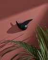Black abstract wall sculpture by Alessi on a textured terracotta wall, with green palm leaves and sharp shadows.