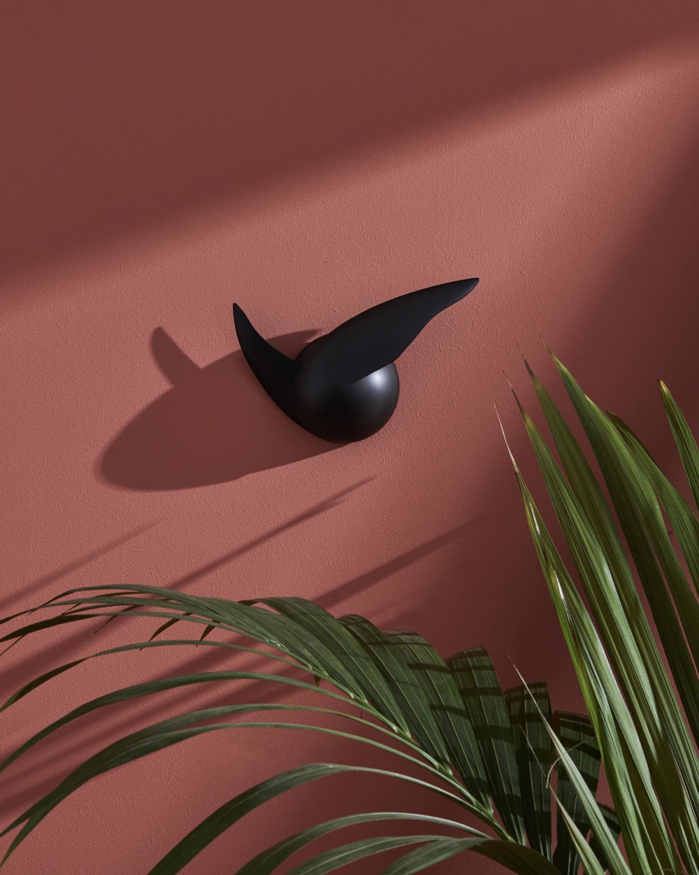 Black abstract wall sculpture by Alessi on a textured terracotta wall, with green palm leaves and sharp shadows.