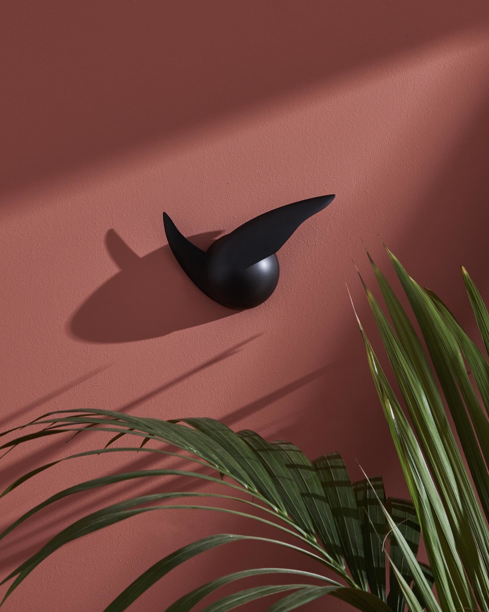 Black abstract wall sculpture by Alessi on a textured terracotta wall, with green palm leaves and sharp shadows.
