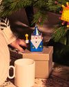Blue and white cube wizard ornament by Alessi, with a starry cone hat, red nose, and star wand, under a Christmas tree.