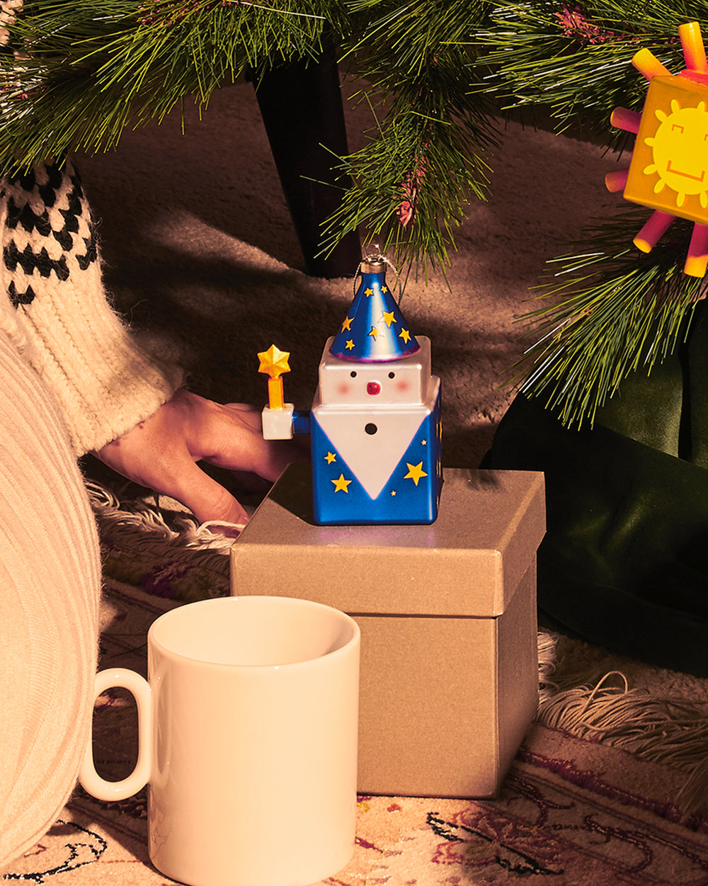 Blue and white cube wizard ornament by Alessi, with a starry cone hat, red nose, and star wand, under a Christmas tree.