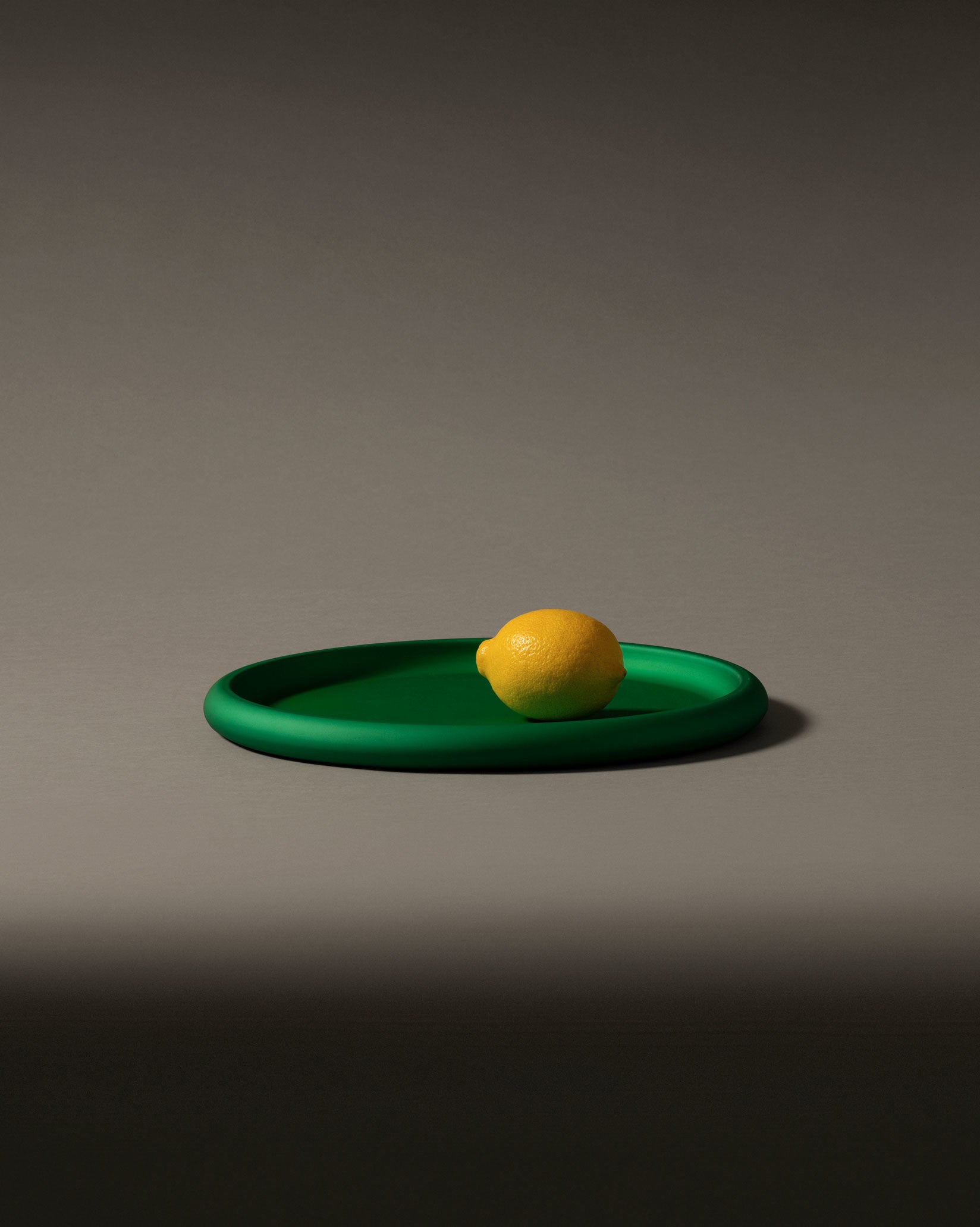 Green matte round fruit bowl by Alessi, holding a vibrant yellow lemon on a neutral background.