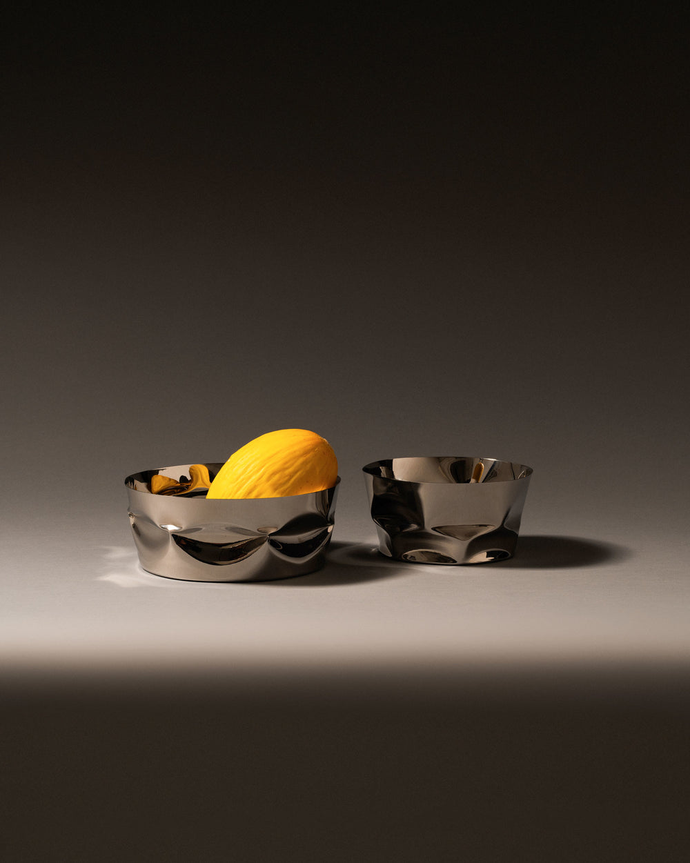 Silver metal fruit bowls by Alessi with a distinct crumpled design, one holding a yellow melon, on a gray surface.
