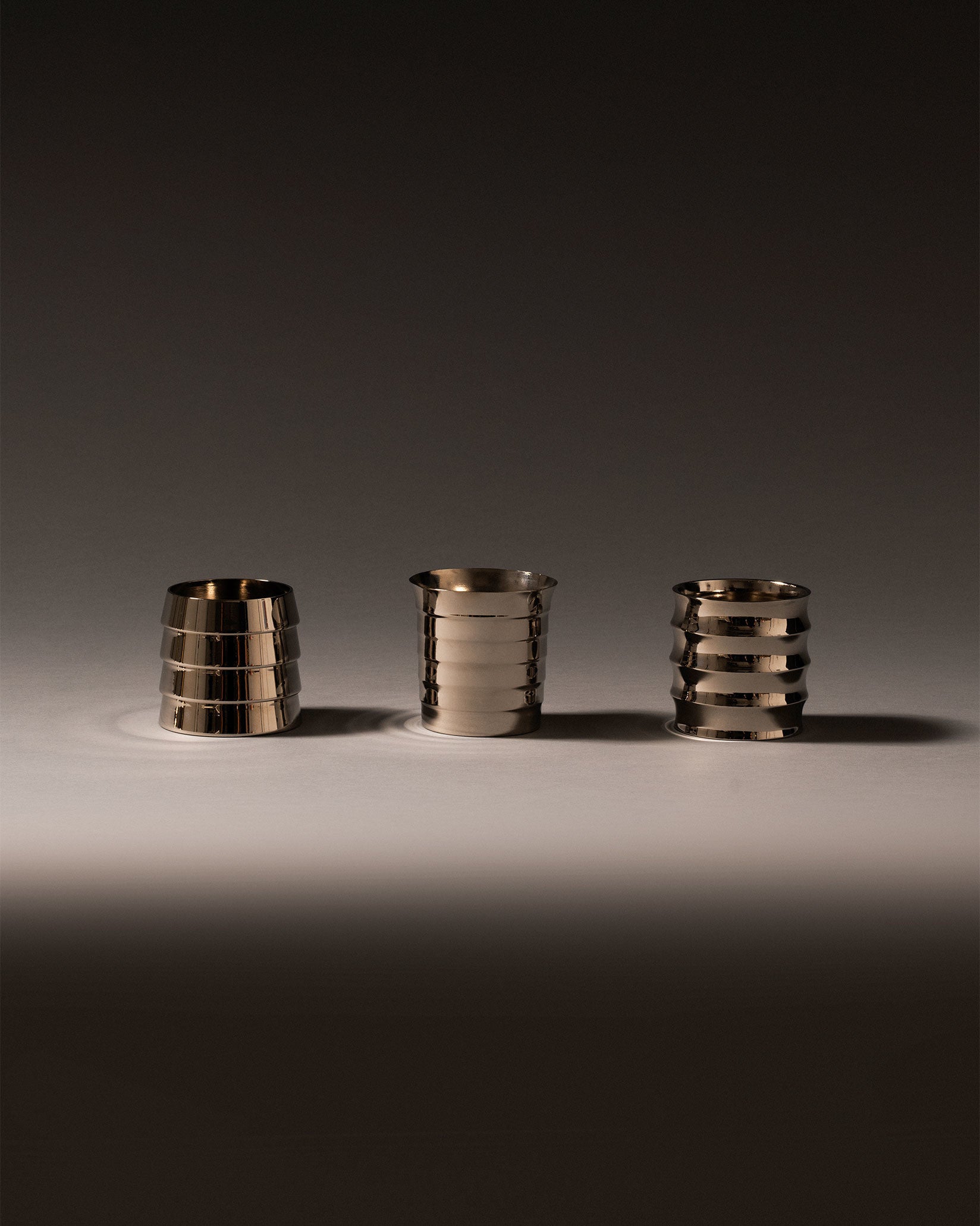 Three metallic silver Dervisci glasses by Alessi with distinct ribbed designs on a light surface.