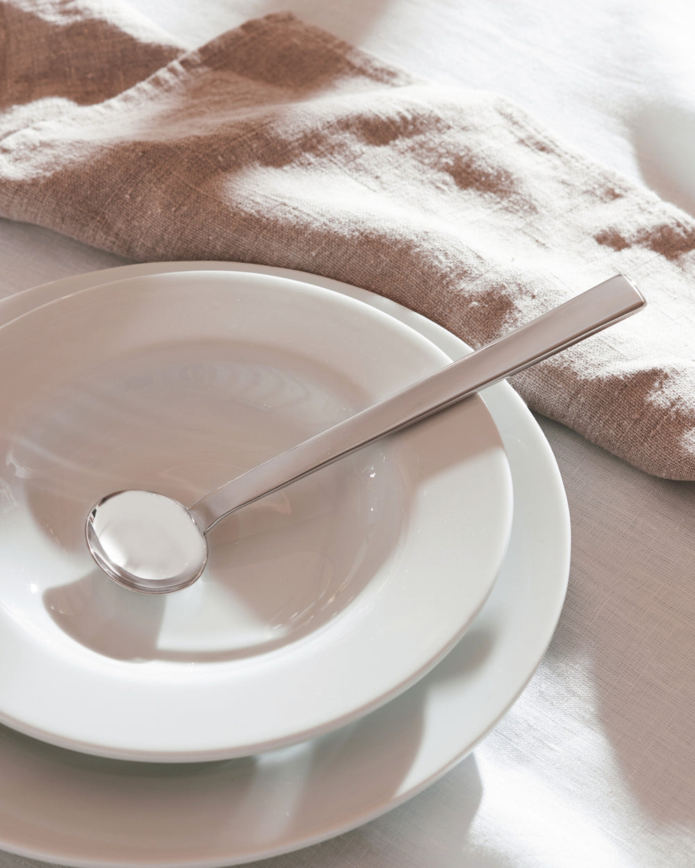 Silver spoon with a round bowl by Alessi resting on white plates and a beige linen napkin.