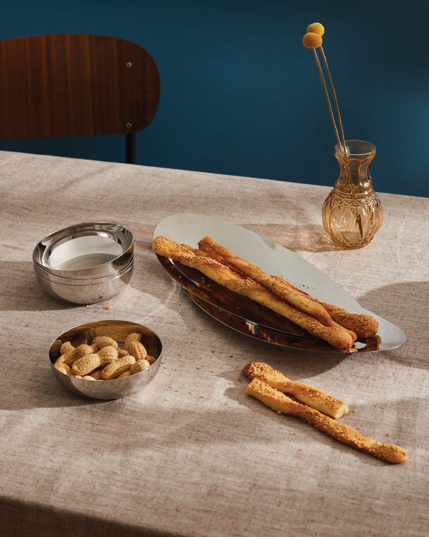 Elongated polished metal Octave basket by Alessi, holding sesame breadsticks on a beige tablecloth.