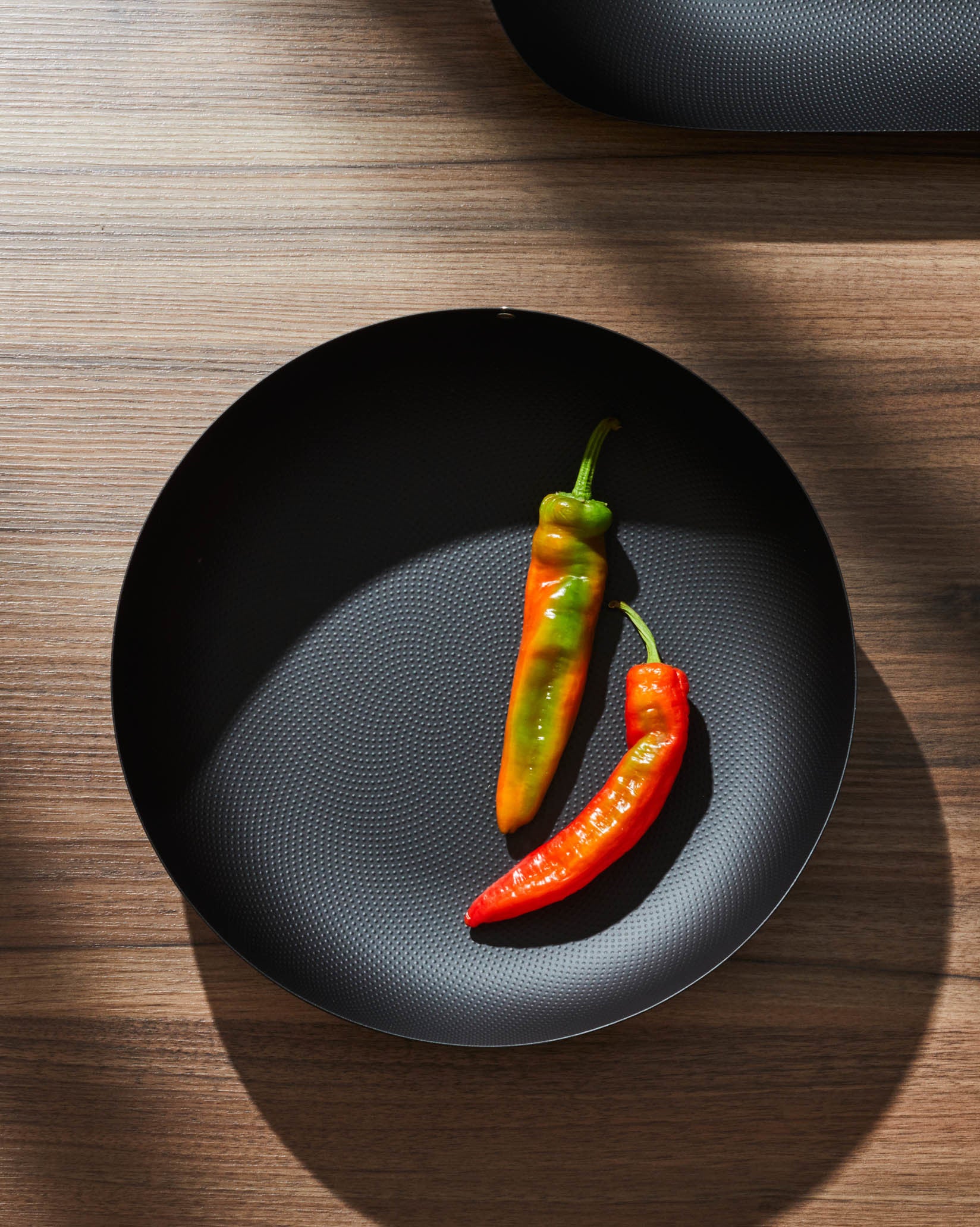 Black round pan by Alessi with dimpled texture, holding two colorful peppers on a wooden table.