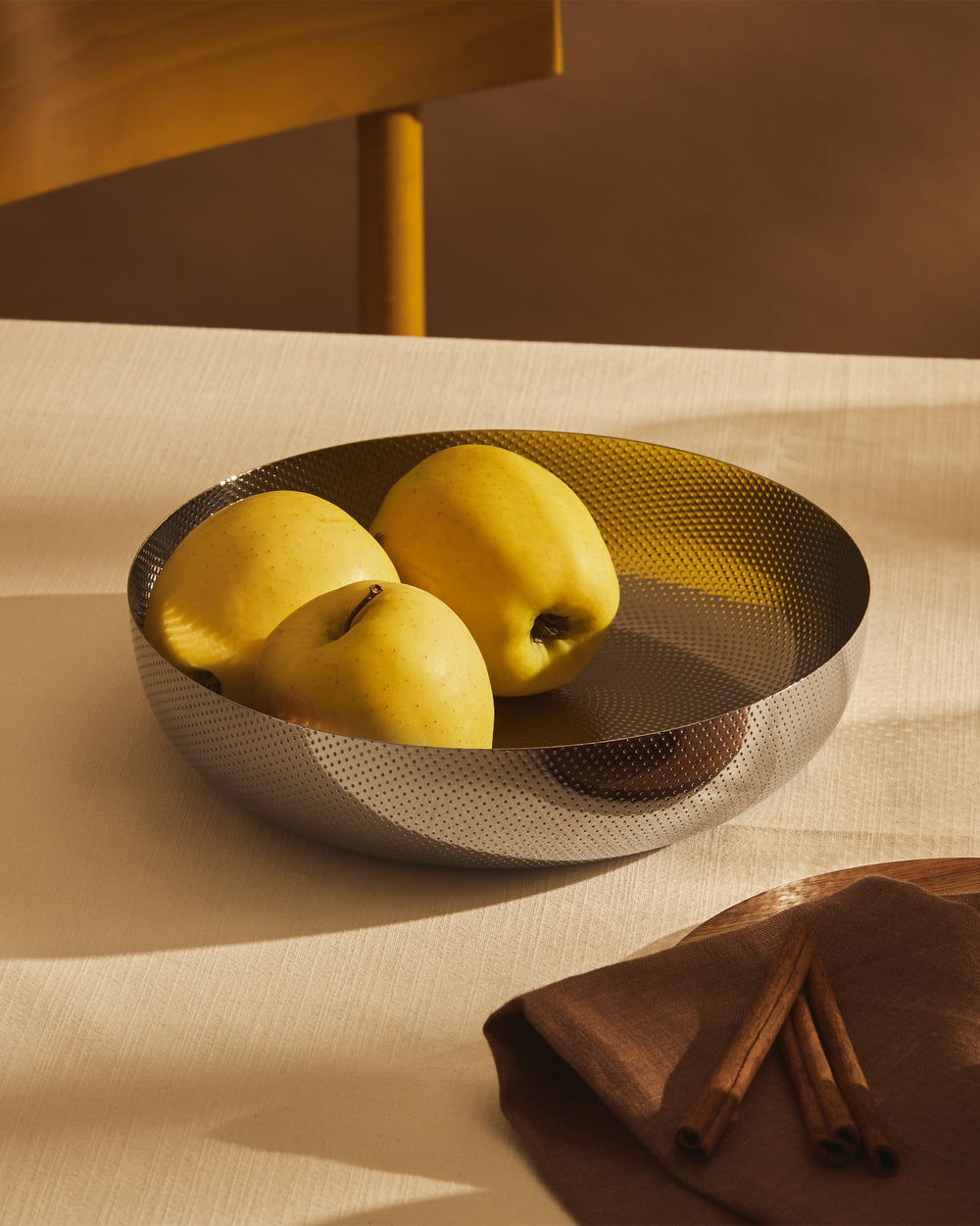 Perforated silver metal bowl by Alessi, containing yellow apples on a light table with cinnamon sticks.