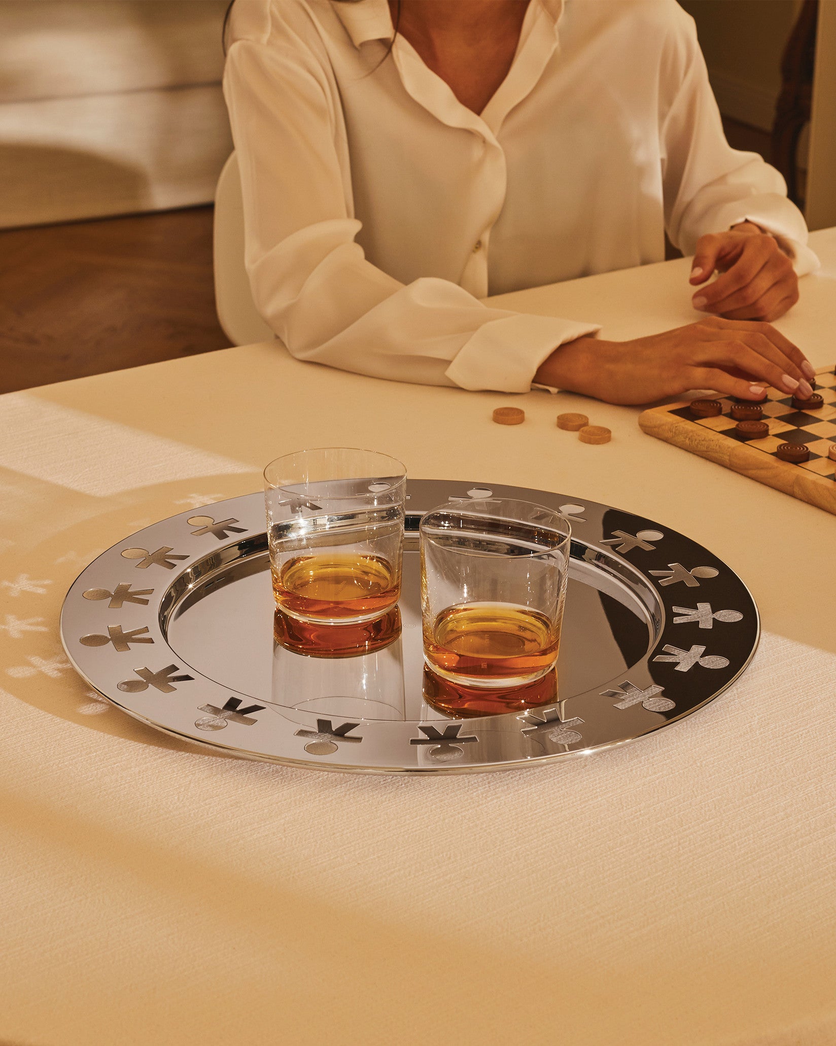 Round polished silver serving tray by Alessi, featuring an edge of cutout "little men" figures, holding two glasses of amber liquid.
