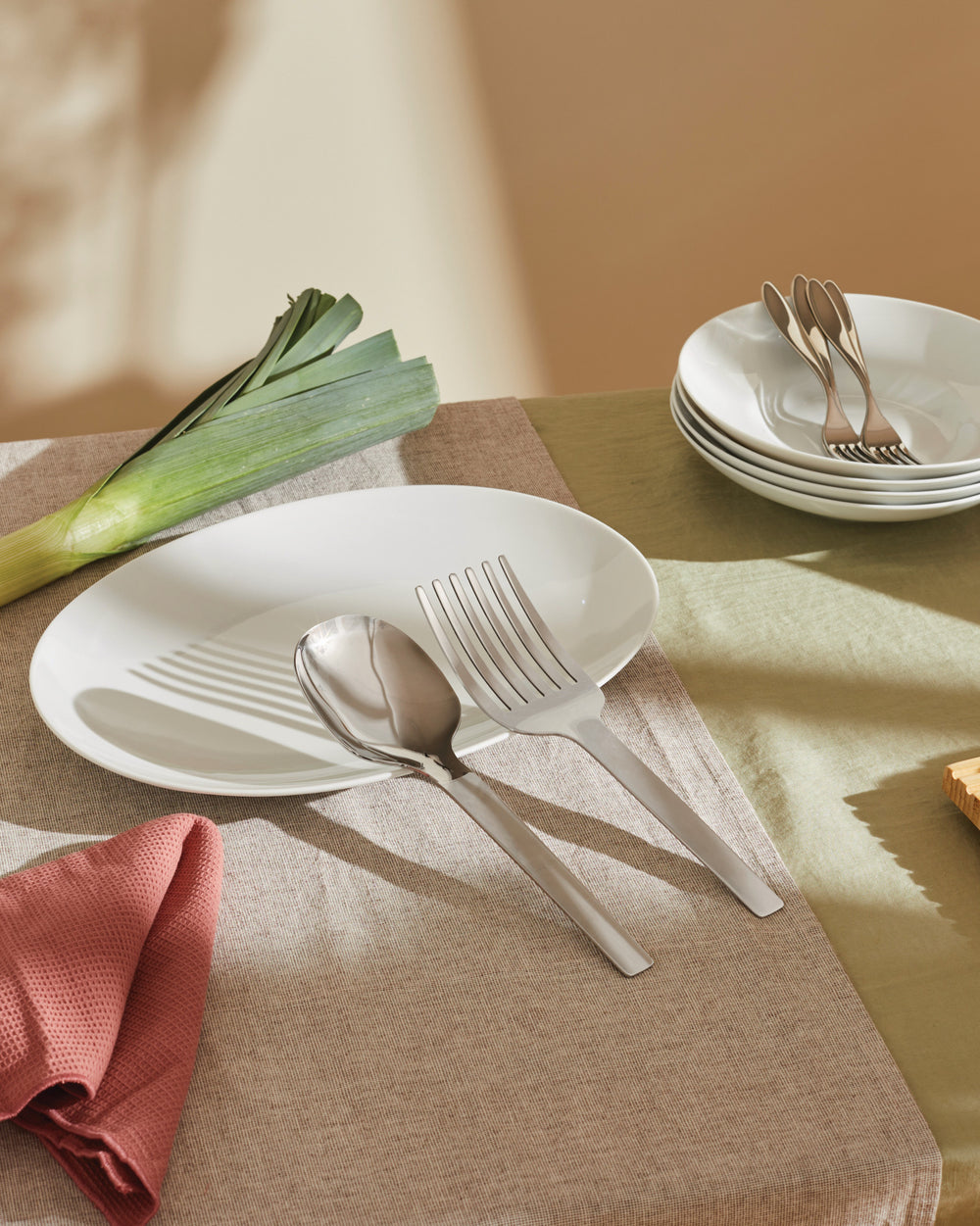 Stainless steel serving cutlery by Alessi, a large spoon and fork with sleek handles on a white plate, alongside a leek and stacked bowls.