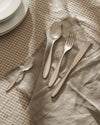 Polished silver Mami flatware set by Alessi, laid on a frayed beige linen napkin over a checkered tablecloth.