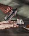 Silver ribbed coffee maker by Alessi, pouring dark coffee into a white cup on a dark table with a pink napkin.