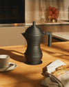 Dark gray ribbed Pulcina coffee maker by Alessi, on a wooden table with a cup and bread.